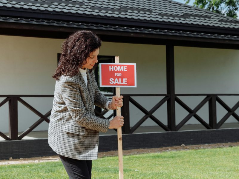 Woman real estate agent placing a 'Home for Sale' sign on a green lawn in front of a house.